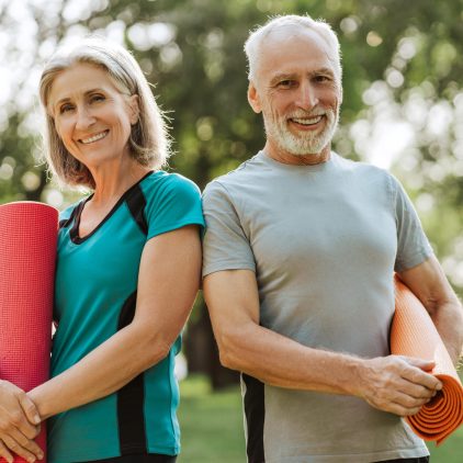 Senior couple in sportswear holds yoga mats in a park, smiling and ready to exercise together. They embody vitality and wellness, finding relaxation and motivation in nature