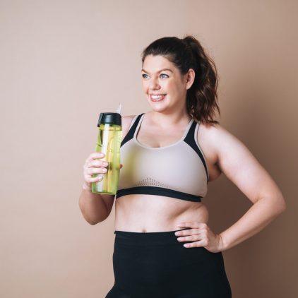 Self loving young woman plus size in sport with bottle of water on beige background, body love